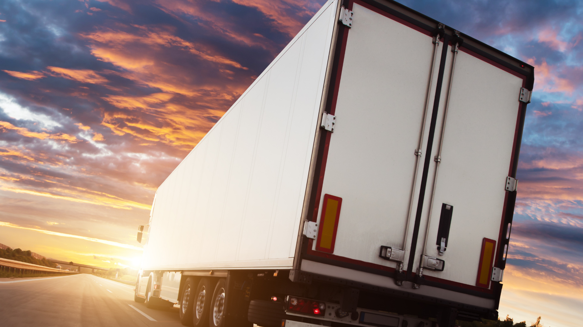 The view from behind a semi truck and trailer on a highway at sunset. 