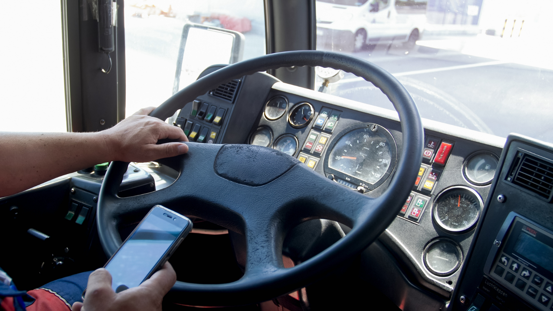 A closeup of a truck driver with one hand on the steering wheel and one hand holding their phone. 