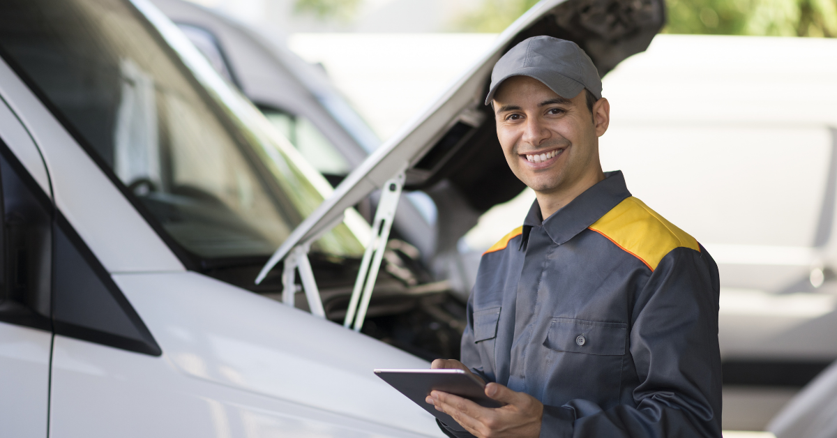 A technician in a uniform smiles and stands beside a white fleet vehicle while using an ELD to check a maintenance issue. 